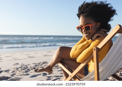 Portrait of african american woman in sunglasses sitting in deckchair smiling on sunny beach by sea. Summer, relaxation, free time and vacation, unaltered. - Powered by Shutterstock