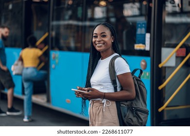 Portrait of an african american woman using smartphone while standing at a bus stop and waiting for public transport at sunny bright day. Black female waiting for a bus. Looking into the camera. - Powered by Shutterstock