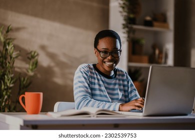 Portrait of an african american woman using laptop while sitting at the table at home and looking into the camera. Young creative black female typing on a keyboard while working remotely. Work at home - Shutterstock ID 2302539753