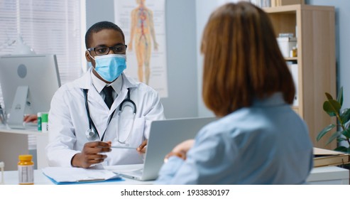 Portrait of African American young male general practitioner in medical mask talking to Caucasian female patient explaining treatment, showing, prescribing pills cure in hospital, medical consultation