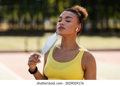 Portrait Of African American Young Woman Taking Off Protective Medical Mask And Breathing Fresh Air, Wearing Wireless Headphones Exercising Outside In The City During Covid Epidemic Outbreak