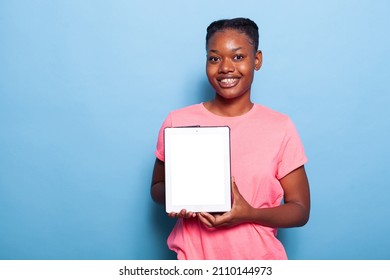 Portrait Of African American Young Woman Holding Tablet Computer With White Screen Standing In Studio With Blue Background. Teenager Recommending Advertisement Ipad Application. Isolated Touchpad