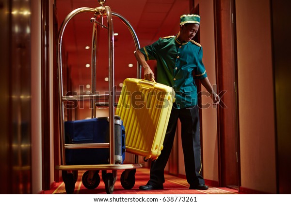 Portrait of African bellboy with\
luggage cart in hotel hallway, bringing bags to guest\
rooms