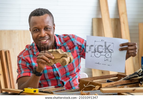 Portrait of African black male carpenter man\
holding wooden toy car model with blueprint in carpentry workshop.\
Concept hobby at home.