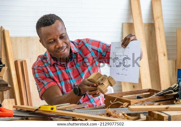 Portrait of African black male carpenter man\
holding wooden toy car model with blueprint in carpentry workshop.\
Concept hobby at home.
