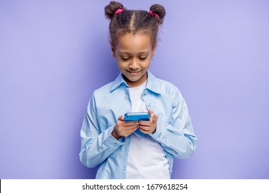 portrait of african child girl with mobile phone in hands, isolated over purple background. sweet child chatting with friend or watching something interesting on phone