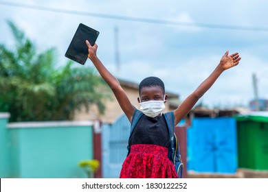 Portrait Of An African Child Student Wearing Face Mask And Holding Digital Tablet With Arms Raised Up - Concept On Education In Covid-19 Pandemic