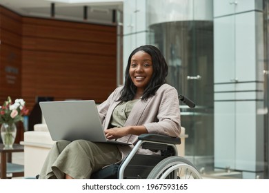Portrait of African disabled woman smiling at camera while sitting in wheelchair and working on laptop