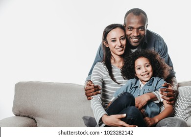 Portrait Of African Family Looking At Camera With Joy While Reposing On Sofa With Coziness. Copy Space In Left Side. Isolated On Background