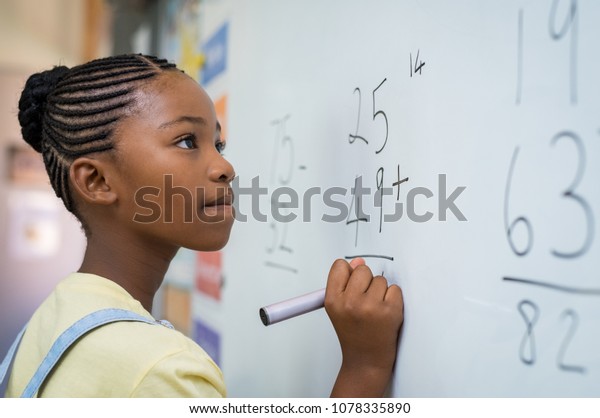 Portrait of african girl writing solution of\
sums on white board at school. Black schoolgirl solving addition\
sum on white board with marker pen. School child thinking while\
doing mathematics\
problem.