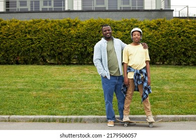 Portrait Of African Happy Boy Standing On Skateboard And Smiling At Camera Together With His Dad While They Riding In Park