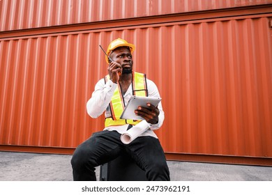 Portrait of an African man engineer or industrial worker in white hard hat, high-visibility Vest, talking on walkie-talkie radio at container yard. Inspector or safety supervisor in container terminal - Powered by Shutterstock