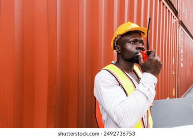 Portrait of an African man engineer or industrial worker in white hard hat, high-visibility Vest, talking on walkie-talkie radio at container yard. Inspector or safety supervisor in container terminal - Powered by Shutterstock