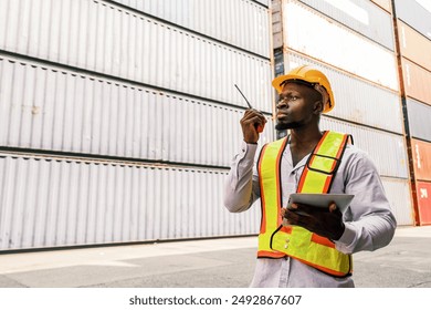 Portrait of an African man engineer or industrial worker in white hard hat, high-visibility Vest, talking on walkie-talkie radio at container yard. Inspector or safety supervisor in container terminal - Powered by Shutterstock