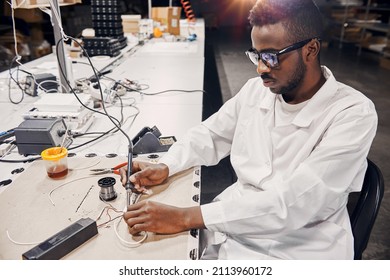 Portrait of african man working with soldering iron, connecting LED lamps and wires. He is wearing white robe, working at the factory
