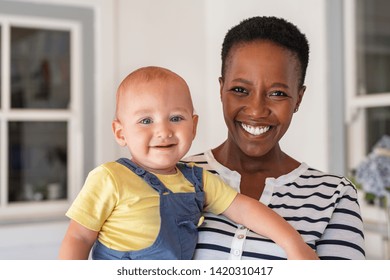Portrait of african mature nanny with baby boy looking at camera. Smiling black mother holding adopted child and smiling together. Portrait of woman relaxing at home with son.