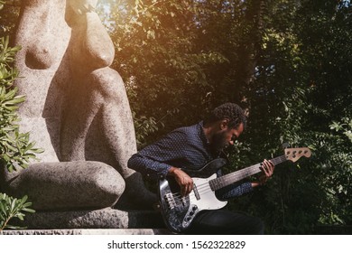 Portrait Of African Musician Bass Guitar Player Outdoors Near The Statue Of A Woman In The Park