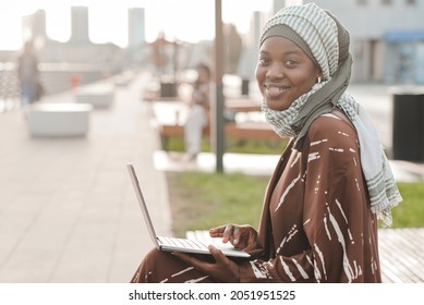 Portrait of African muslim woman smiling at camera while sitting on the bench and using laptop outdoors - Shutterstock ID 2051951525