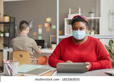 Portrait Of African Overweight Woman In Protective Mask Looking At Camera While Sitting At Workplace And Working On Tablet Pc At Office