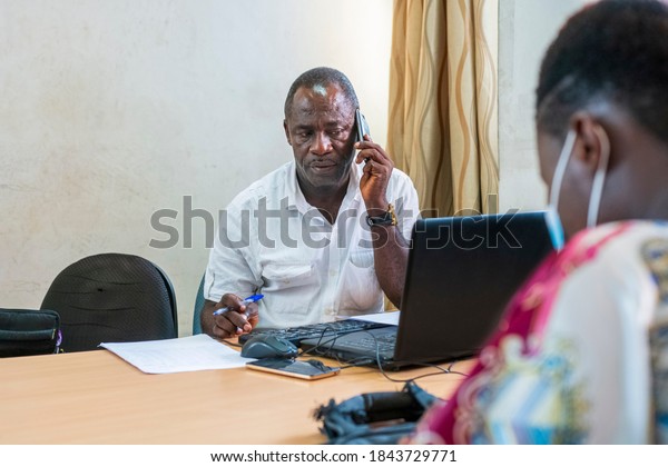 portrait of an African senior\
citizen or old man sitting in office with cell phone attached to\
left ear
