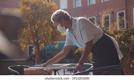 Portrait Of African Waitress Wearing Face Mask Cleaning Table Outdoors. Young African Woman Small Business Owner Wiping Table With Cloth On Restaurant Terrace