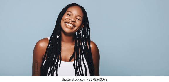 Portrait of an African woman with dreadlocks and body piercings smiling at the camera. Happy young woman feeling confident in her style. Fashionable woman standing against a studio background. - Shutterstock ID 2278540099