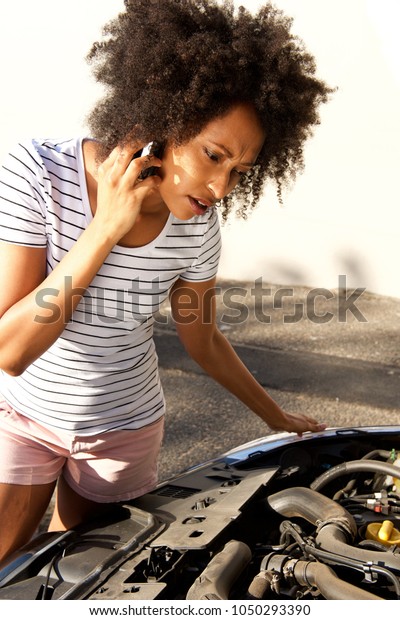 Portrait of african woman looking at her\
broken car and making phone call for\
assistance