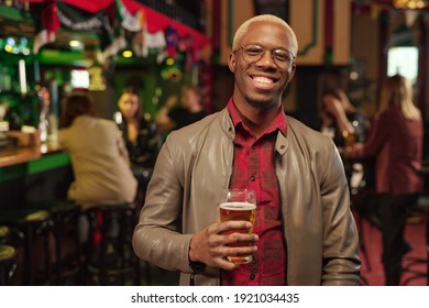 Portrait of African young man holding glass of beer and smiling at camera while standing in the bar