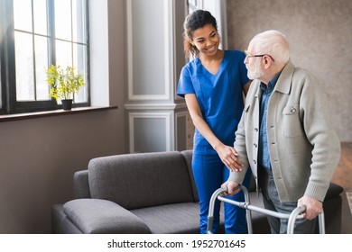 Portrait of an african young nurse helping old elderly disable man grandfather to walk using walker equipment in the bedroom. Senior patient of nursing home moving with walking frame and nurse support