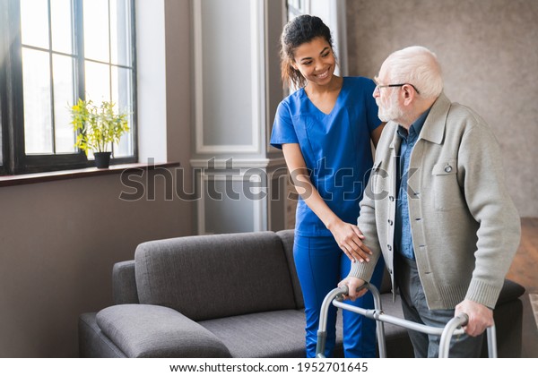 Portrait of an african young nurse helping old\
elderly disable man grandfather to walk using walker equipment in\
the bedroom. Senior patient of nursing home moving with walking\
frame and nurse\
support