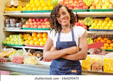 Portrait Of African Young Woman In Jeans Apron Standing With Arms Crossed And Smiling At Camera With Shelves Of Fruits And Vegetables In The Background In The Market