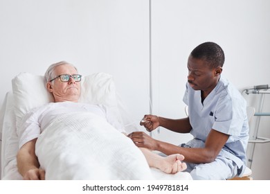 Portrait Of African-American Male Nurse Caring For Senior Man Lying In Hospital Bed Againts White, Copy Space