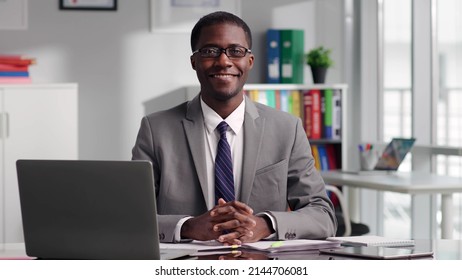 Portrait Of African-American Politician In Glasses Smiling At Camera Sitting At Desk At Modern Spacious Workplace