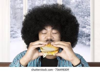 Portrait Of Afro Man With Curly Hair, Eating Hamburger At Home With Winter Background On The Window