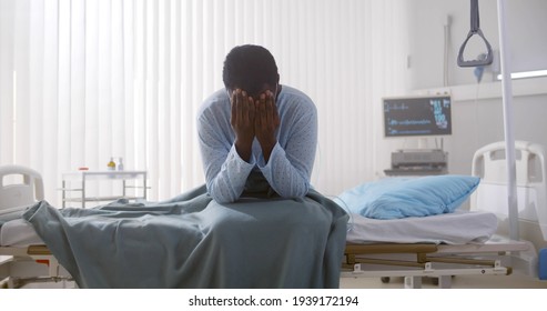 Portrait Of Afro-american Sick Patient Sitting On Hospital Bed And Rubbing Face Feeling Unwell. Tired And Exhausted Sick Black Man Sitting In Hospital Ward