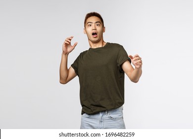 Portrait of alarmed and scared, shocked asian guy gasping, step back and raising hands up frightened, startled staring at something frightening, standing in awe over grey background