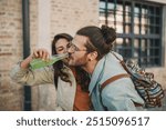 Portrait of alternative hipster girl giving her boyfriend bottle of drink on a city street at old town. Young silly trendy urban guy drinking bottle of cold fresh beverage on a city street downtown.