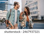 Portrait of alternative young trendy tourist couple walking on urban city street with skateboards in hands and smiling at each other. Hipster visitors talking and walking on city street downtown.