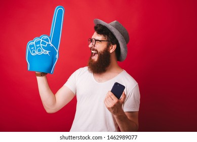 Portrait Of Amazed Bearded Man Celebrating Success Holding Smartphone And Foam Finger 