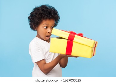 Portrait Of Amazed Curious Adorable Little Boy Peeking Inside Gift Box, Unpacking Present With Funny Astonished Expression, Impatient Child Unboxing Birthday Surprise. Studio Shot Blue Background