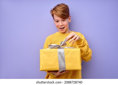 Portrait of amazed curious adorable little boy unpacking present box with funny astonished expression, impatient caucasian child unboxing birthday surprise. studio shot purple background.