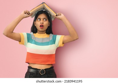 Portrait of an amazed happy indian asian girl student with open mouth and big eyes posing isolated holding book on her head and looking away at copy space.