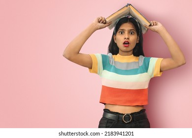 Portrait of an amazed indian asian girl student with open mouth and big eyes posing isolated holding book on her head and looking directly to camera with copy space.