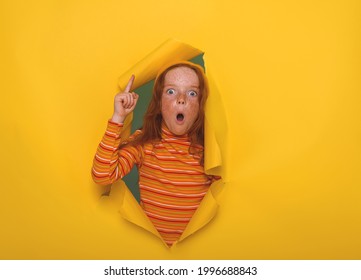 Portrait Of Amazed Or Surprised Freckled Redhead Child Girl Breaking Through Yellow Paper Wall. Kid Pointing Up With Finger. Sale Discount. Funny Face With Open Mouth And Big Eyes.