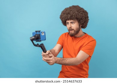 Portrait of amazed surprised man with Afro hairstyle standing with steadicam and having video call, looking at phone camera with excitement. Indoor studio shot isolated on blue background. - Powered by Shutterstock