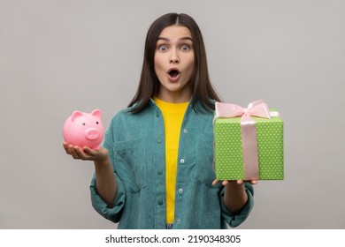 Portrait of amazed surprised woman holding wrapped gift box and piggy bank, save money, looking at camera with open mouth, wearing casual style jacket. Indoor studio shot isolated on gray background.