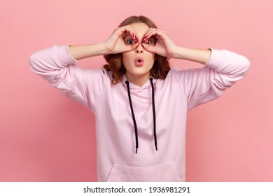 Portrait of amazed teen girl with curly hair in hoodie looking through binoculars gesture and expressing surprise, zooming vision, exploring distance. Indoor studio shot isolated on pink background