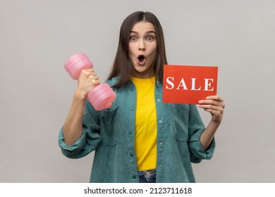 Portrait Amazed Woman Showing Holding Sale Card And Dumbbell, Black Friday Discounts On Gym Membership, Wearing Casual Style Jacket. Indoor Studio Shot Isolated On Gray Background.