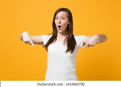 Portrait Of Amazed Young Woman In White Casual Clothes Keeping Mouth Open Pointing Index Fingers Down Isolated On Yellow Orange Wall Background In Studio. People Lifestyle Concept. Mock Up Copy Space