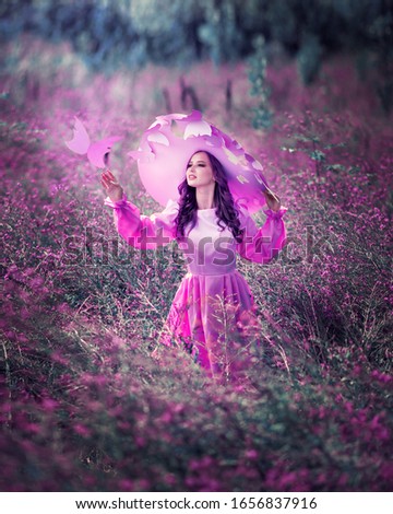 Portrait of amazing cute young woman in beautiful fairy tale image in purple fluffy long dress and with big hat stands in field with purple flowers and stretches her hand to paper purple butterfly